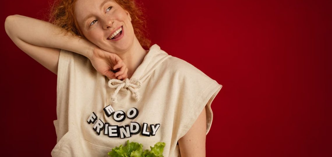Cheerful woman with red hair in eco-friendly attire holding fresh lettuce against a vibrant red background.
