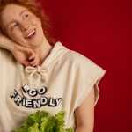 Cheerful woman with red hair in eco-friendly attire holding fresh lettuce against a vibrant red background.