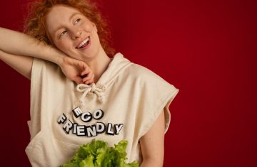 Cheerful woman with red hair in eco-friendly attire holding fresh lettuce against a vibrant red background.