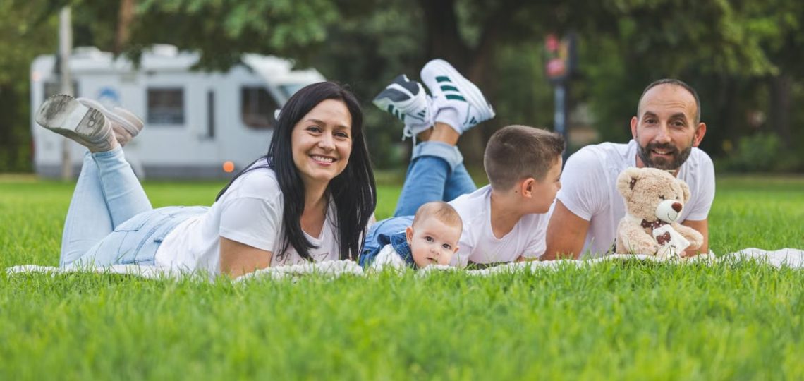 Happy family lying on grass with RV in the background, enjoying a sunny outdoor picnic.