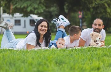 Happy family lying on grass with RV in the background, enjoying a sunny outdoor picnic.