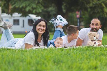Happy family lying on grass with RV in the background, enjoying a sunny outdoor picnic.